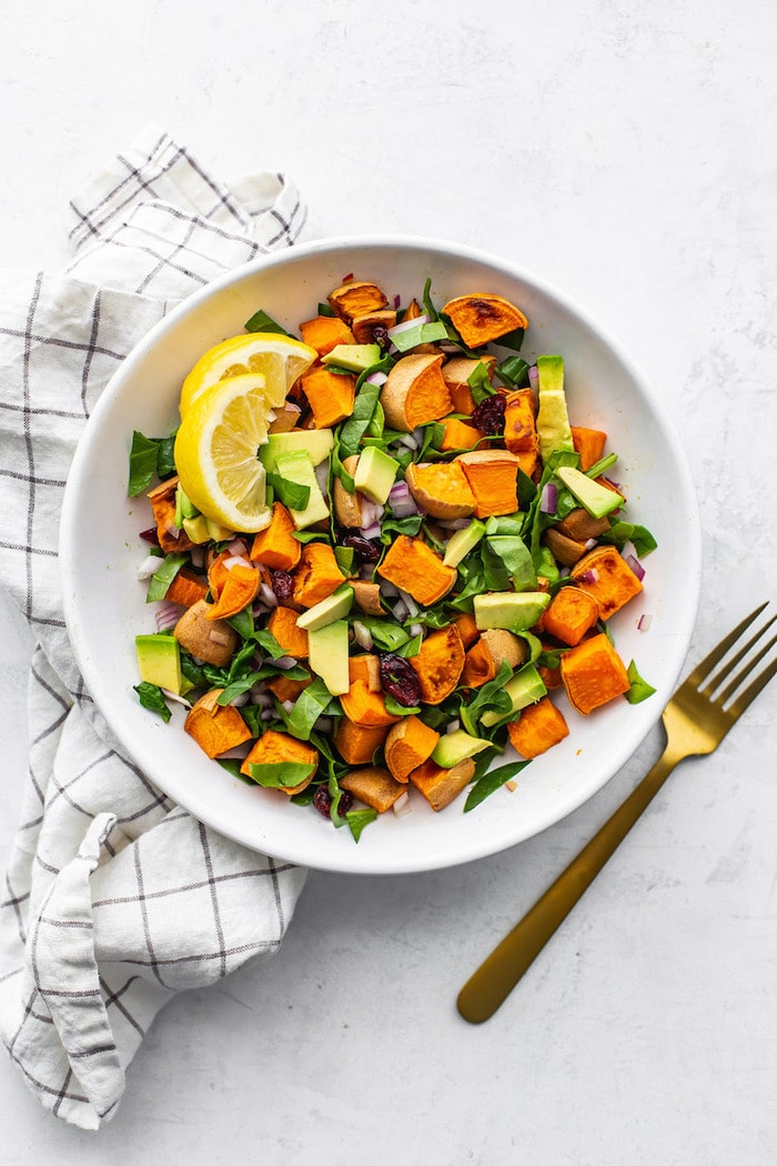 Bowl of sweet potato, avocado and spinach salad garnished with lemon wedges. A fork and napkin are beside the bowl.