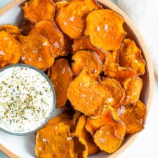 Baked homemade sweet potato chips on a bowl and topped with flakey salt. A bowl of herby creamy dip is beside the chips.