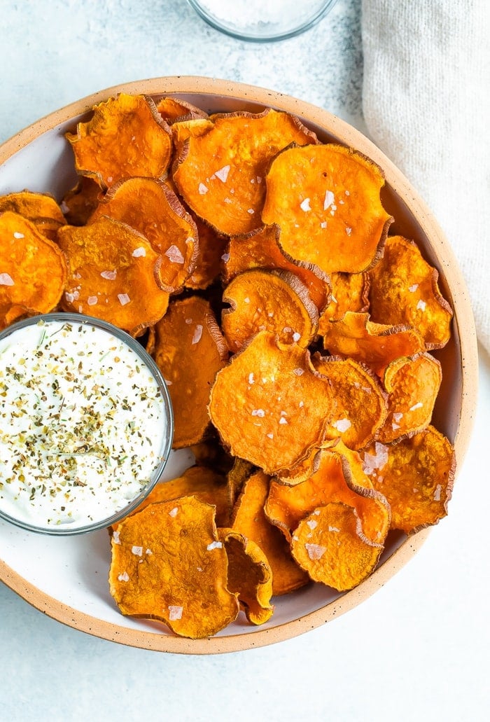 Baked homemade sweet potato chips on a bowl and topped with flakey salt. A bowl of herby creamy dip is beside the chips.