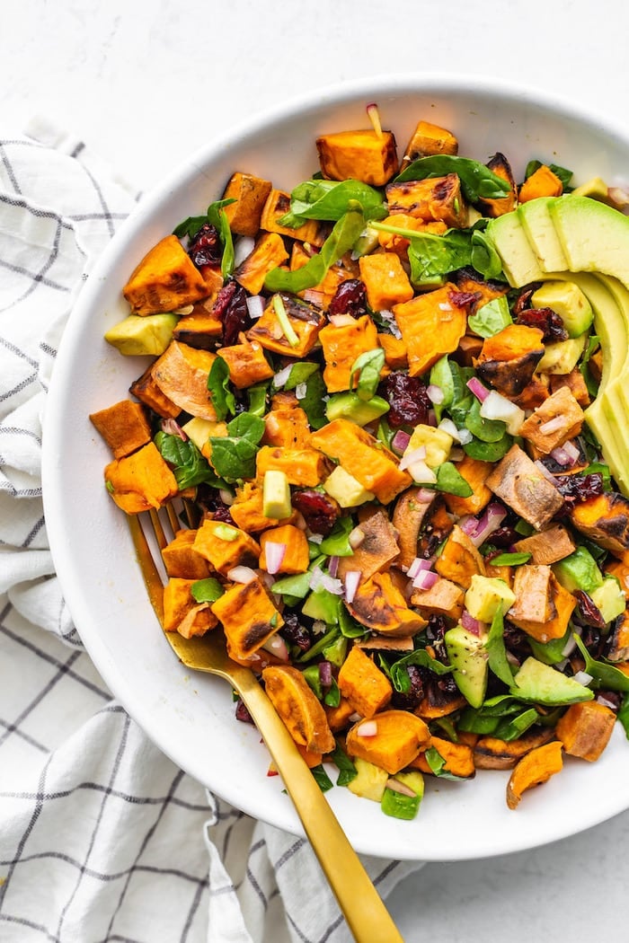 Bowl of sweet potato salad with avocado, spinach, onion and cranberries.