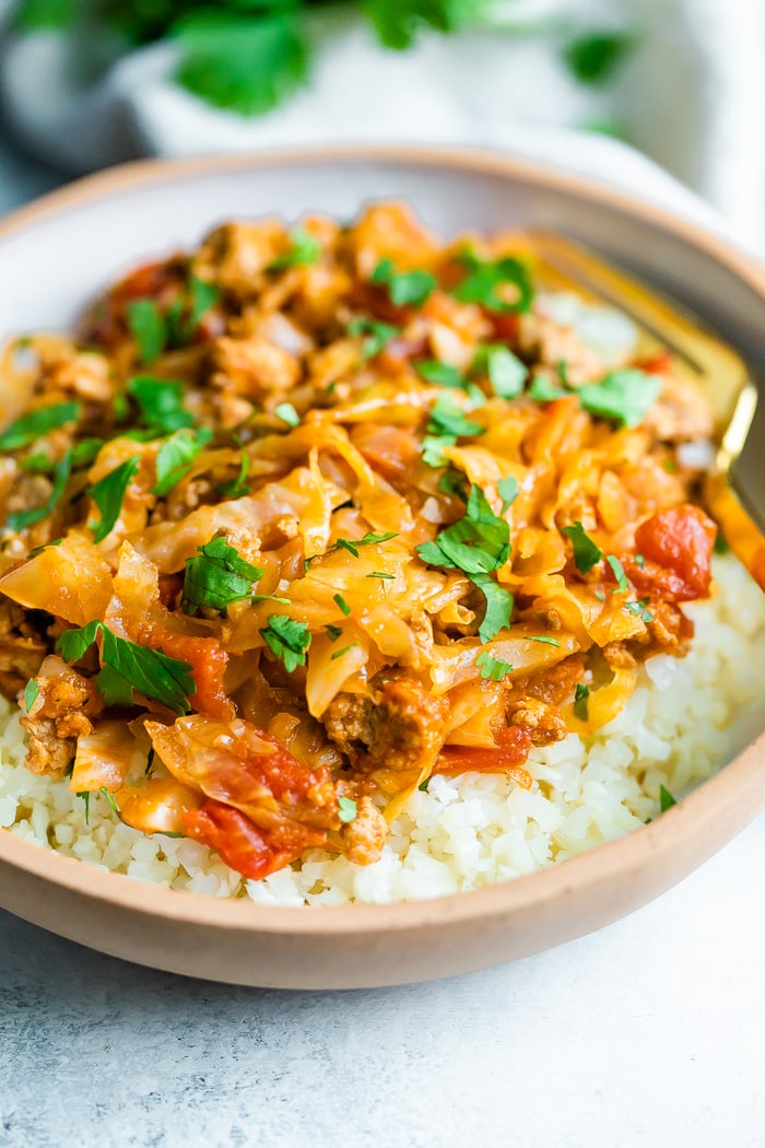 Close up image of unstuffed cabbage roll bowl. Cabbage and turkey is mixed with a tomato sauce and served over rice. Food is served in a ceramic bowl with a gold fork resting inside the bowl.