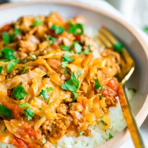 Close up shot of unstuffed cabbage roll bowl. Cabbage and turkey are mixed with a tomato sauce and served over rice. Gold fork is resting inside white ceramic bowl.