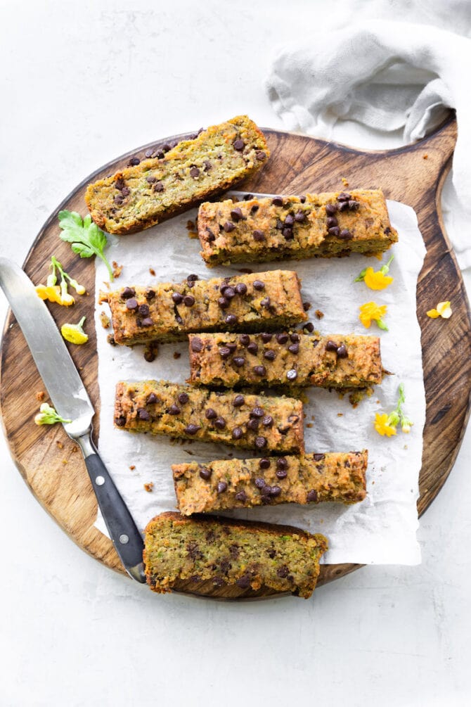 Slices of zucchini bread on a wooden cutting board surrounded by yellow flowers, a cloth napkin and a knife.