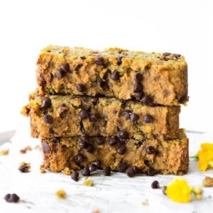 Three slices of chocolate chip zucchini bread stacked on top of each other. Chocolate chips and yellow flowers are scattered around.