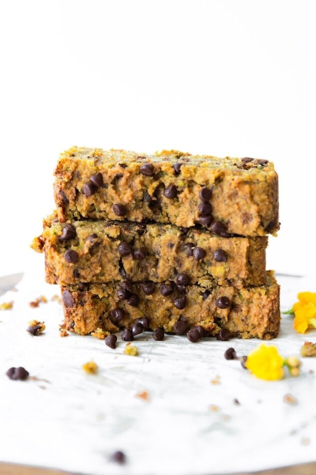 Three slices of chocolate chip zucchini bread stacked on top of each other. Chocolate chips and yellow flowers are scattered around.