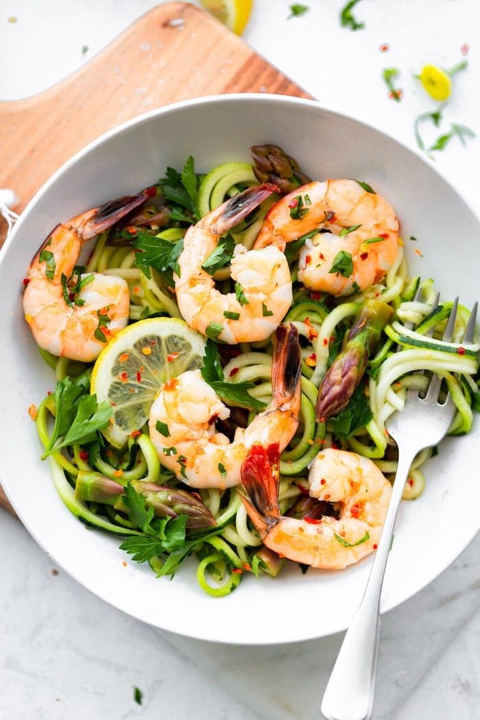 Bowl with zucchini noodles, asparagus and shrimp garnished with lemon and red pepper flakes.