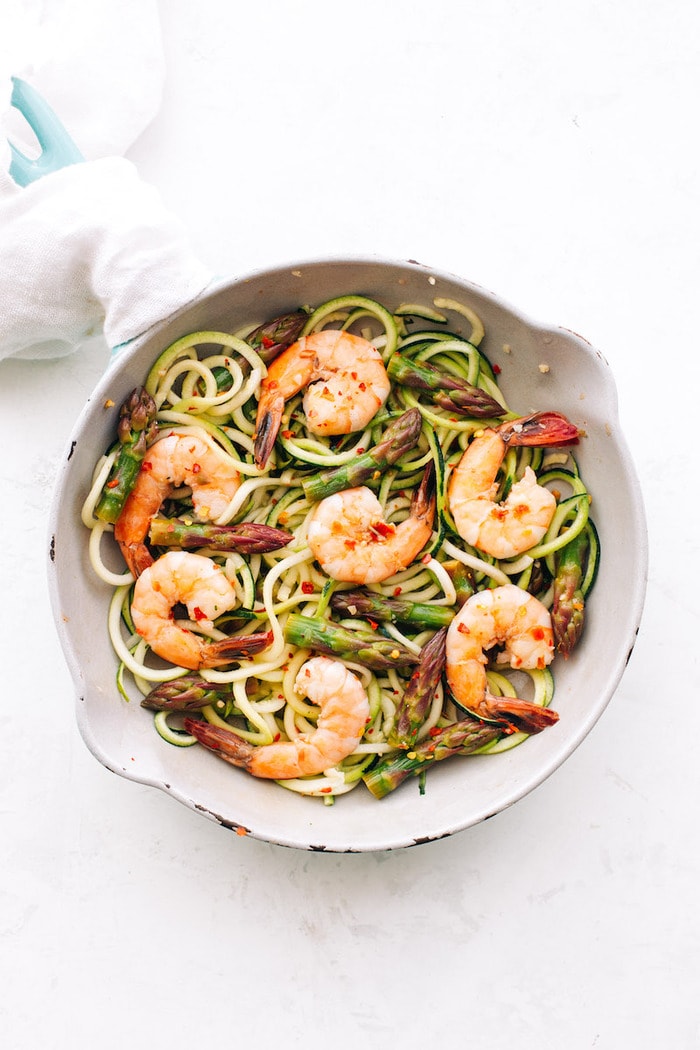 White sauté pan with zucchini noodles, asparagus and shrimp topped with red pepper flakes.