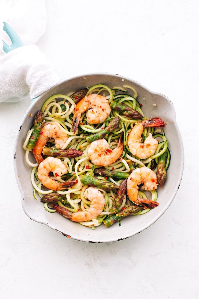 White sauté pan with zucchini noodles, asparagus and shrimp topped with red pepper flakes.