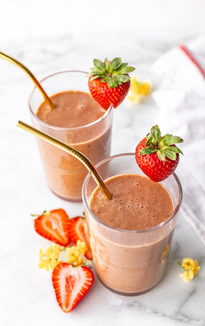 Two glasses with metal straws and full of chocolate strawberry smoothies. The glasses are garnished with a strawberry. Strawberries and flowers are around the glasses.