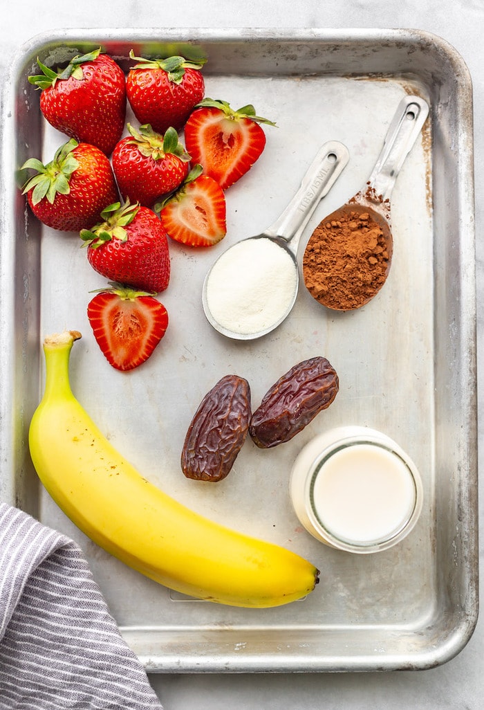 Ingredients to make a chocolate strawberry smoothie on a cookie sheet. Ingredients on the cookie sheet are strawberries, protein powder, cocoa powder, dates, almond milk and banana.