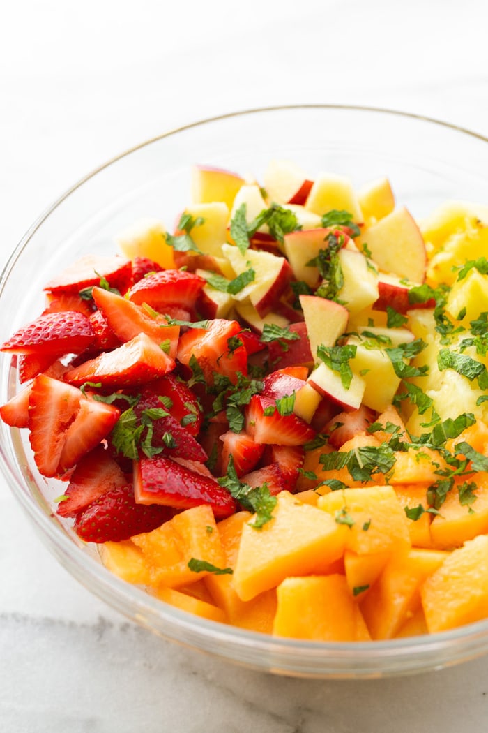 Glass mixing bowl with chopped strawberries, melon, apples, pineapple and fresh mint.