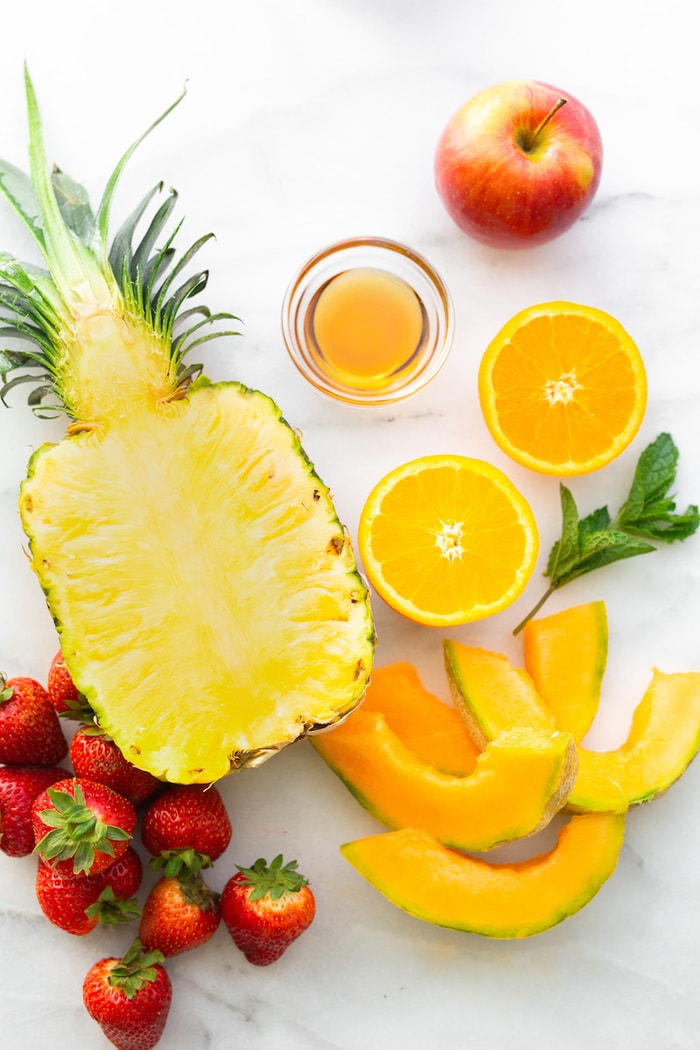 Marble counter with a bowl of maple, sliced-in-half oranges, a pineapple, strawberries, melon, an apple and a spring of fresh mint.