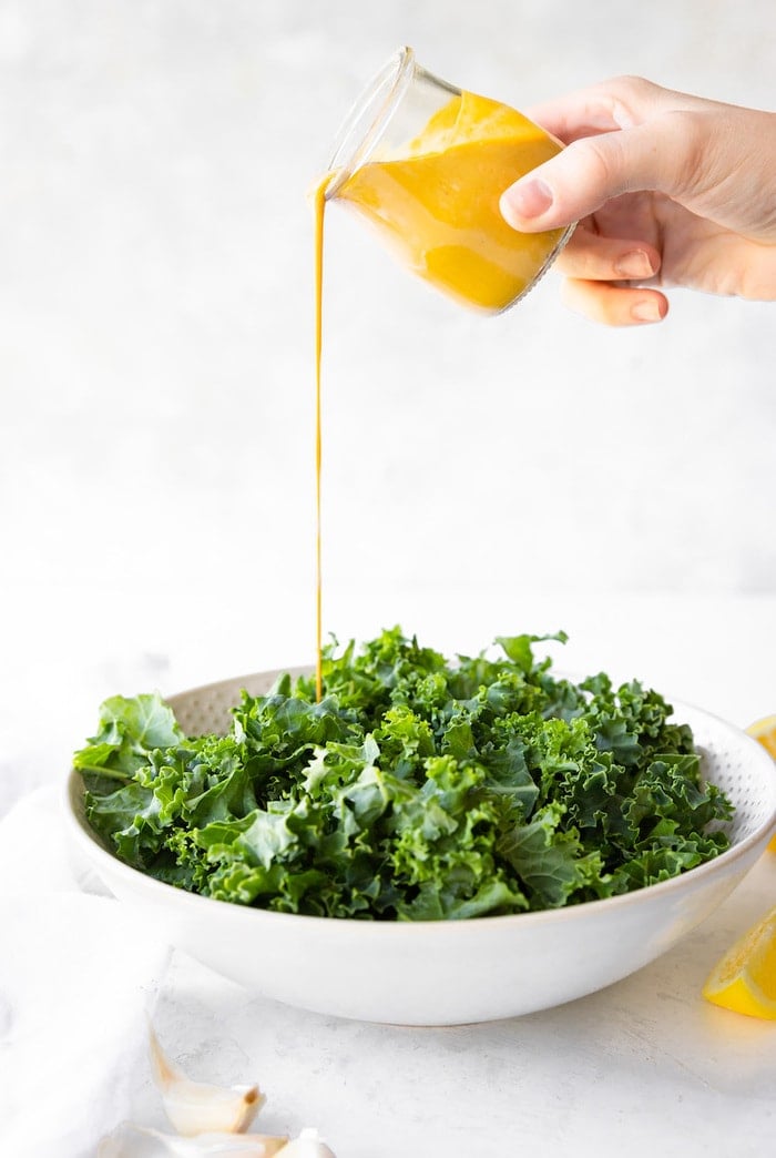 Raw kale in a white bowl with tahini dressing being poured on.
