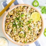 Bird's eye view photo of a bowl of Mexican Street Corn Salad topped with lime and fresh cilantro. Bowl is resting on a striped napkin and next to a gold spoon and bowl of cheese.