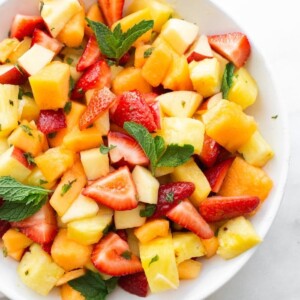 White bowl with fruit salad including strawberries, pineapple, melon, apples and mint.