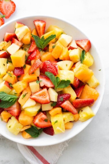 White bowl with fruit salad including strawberries, pineapple, melon, apples and mint.