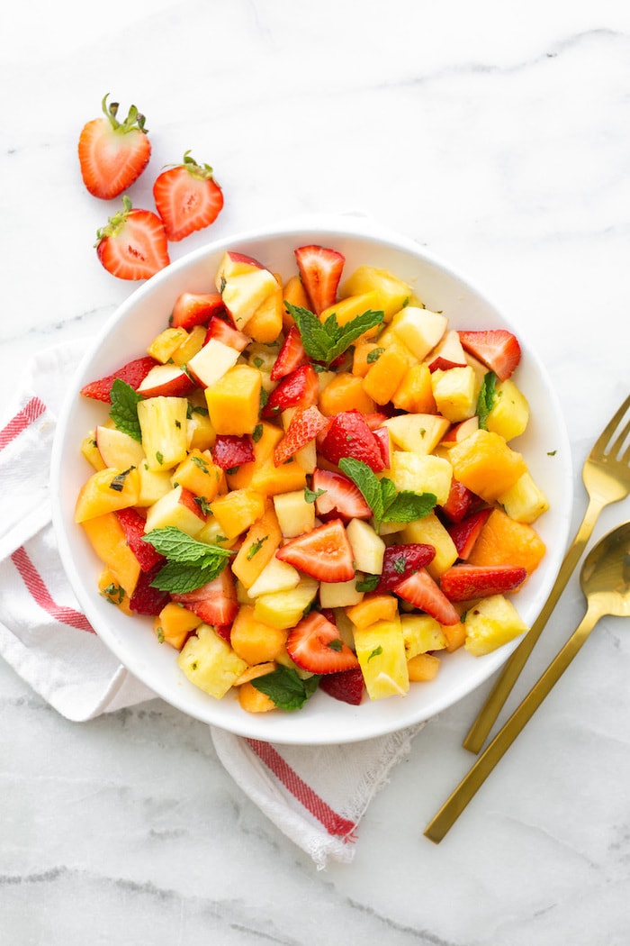 White bowl with fruit salad including strawberries, pineapple, melon, apples and mint. A napkin and serving utensils are beside the bowl.