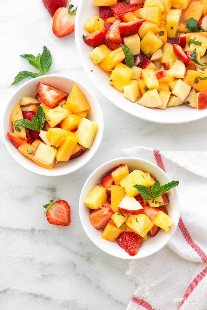 Large serving bowl and two small bowls filled with fruit salad garnished with fresh mint.