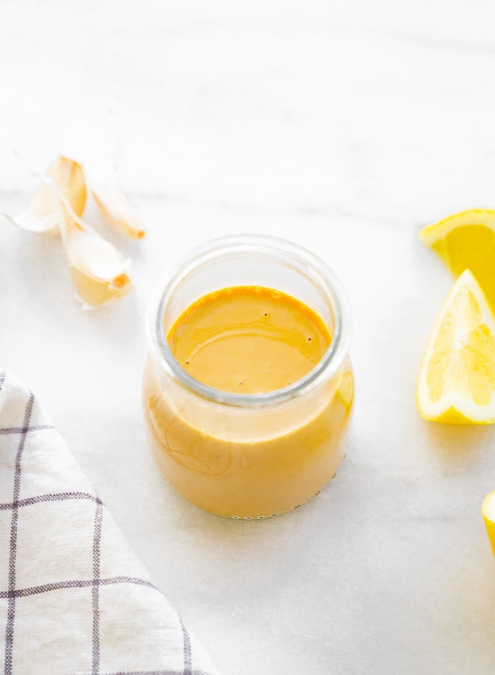 Zesty tahini salad dressing in a glass jar with garlic, lemon and a towel on the side.