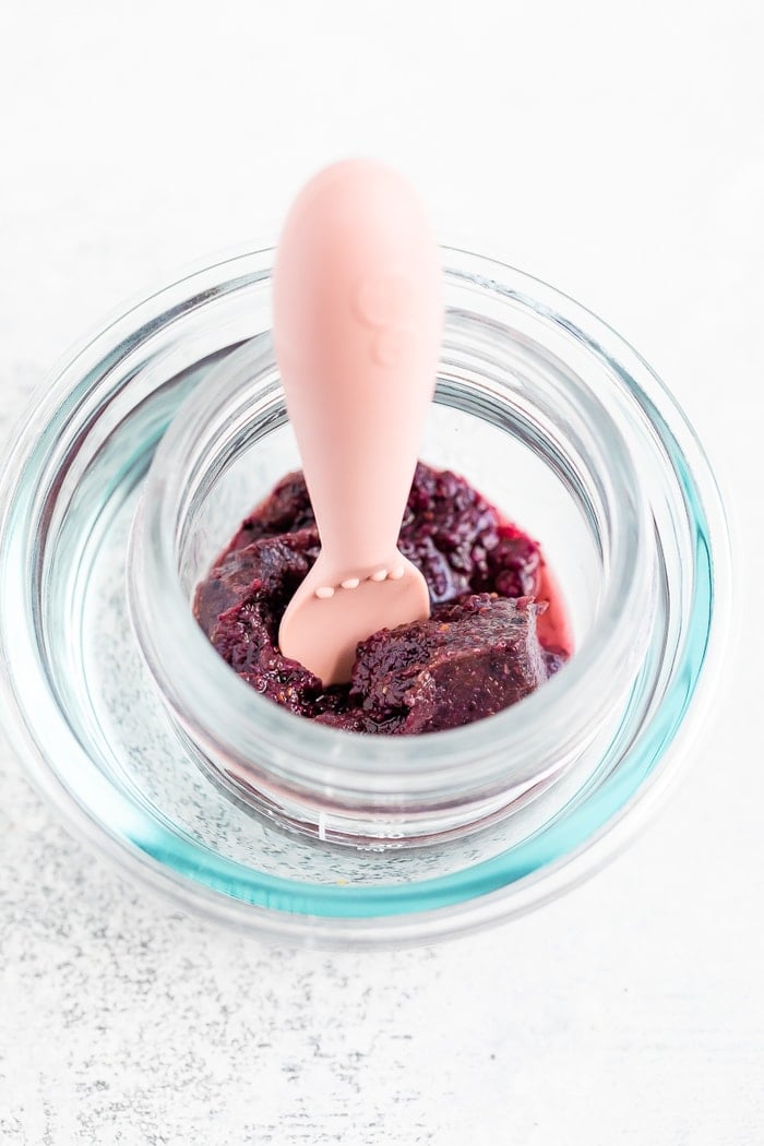 Frozen blueberry baby food cube in a water bath to thaw with pink baby spoon.