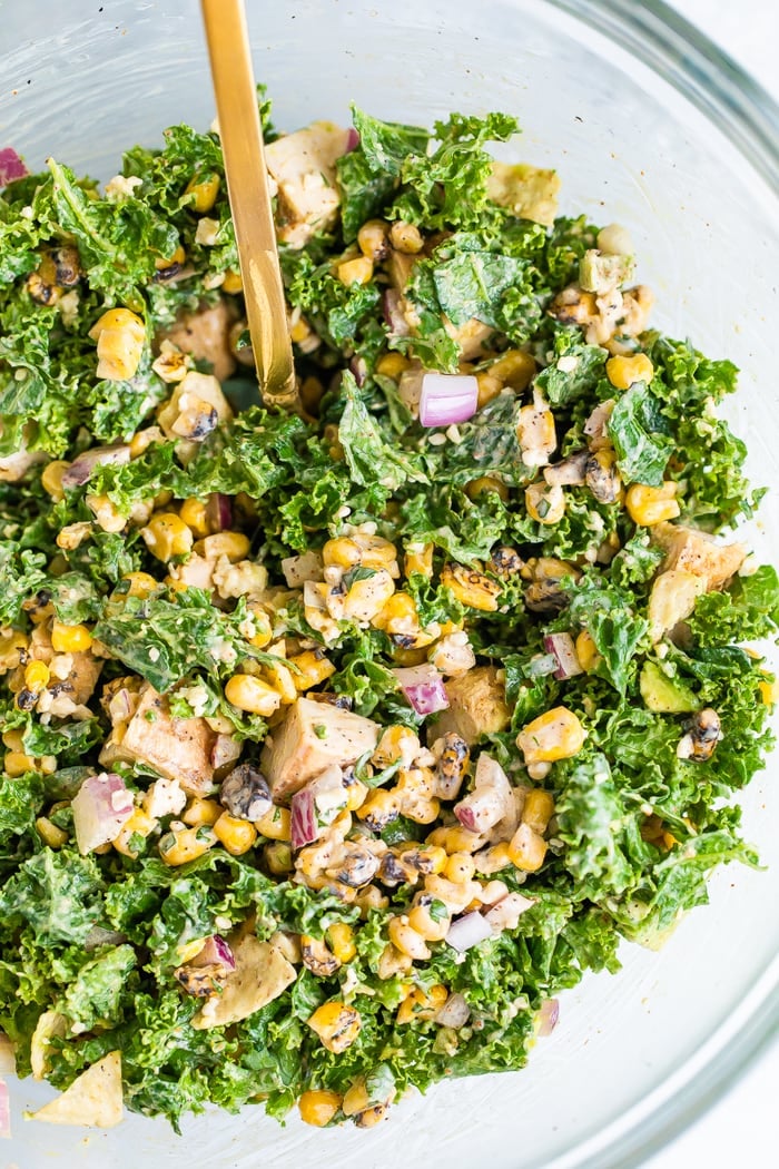 Glass mixing bowl with a gold spoon in a kale salad made with Mexican street corn, grilled chicken, avocado and crushed tortilla chips.