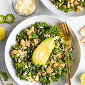 Close up photo of a white bowl and gold fork with a Mexican street corn and kale grilled chicken salad. The salad is topped with slices of avocado. Ingredients in the salad are sprinkled on the table.