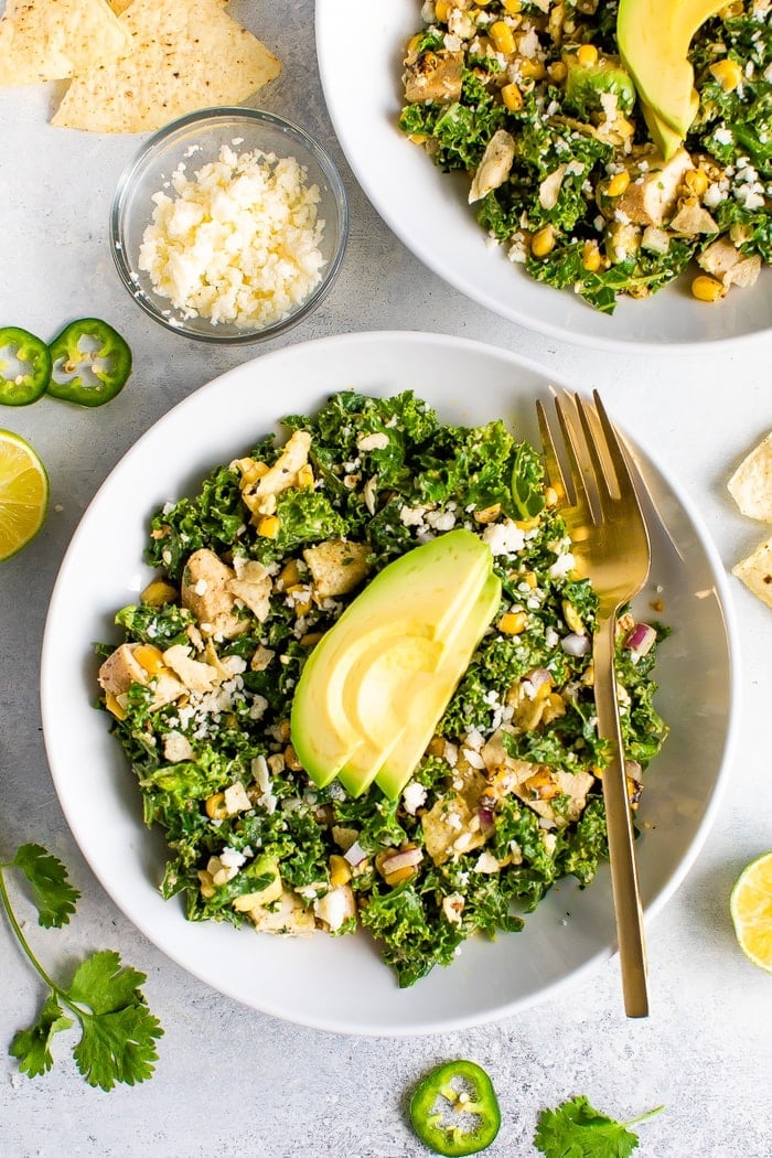 Close up photo of a white bowl and gold fork with a Mexican street corn and kale grilled chicken salad. The salad is topped with slices of avocado. Ingredients in the salad are sprinkled on the table.