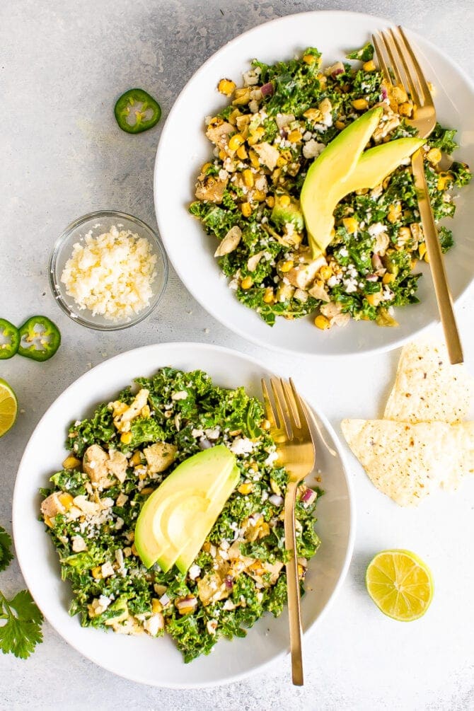 Two white bowls filled with kale Mexican street corn salad with grilled chicken and topped with slices of avocado. Two gold forks are resting on the side of the plates. On the table, cilantro, a bowl of cheese, slices of jalapeno and lime and tortilla chips are scattered.