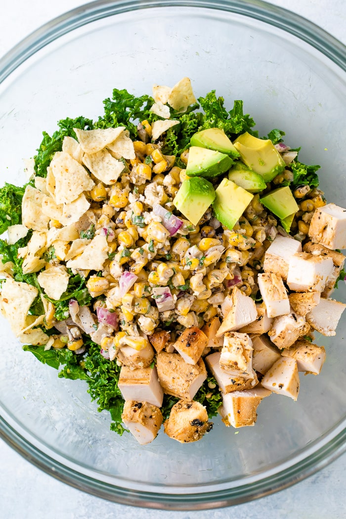 Glass mixing bowl with ingredients for a salad including kale, Mexican street corn salad, crushed tortilla chips, avocado and grilled chicken.