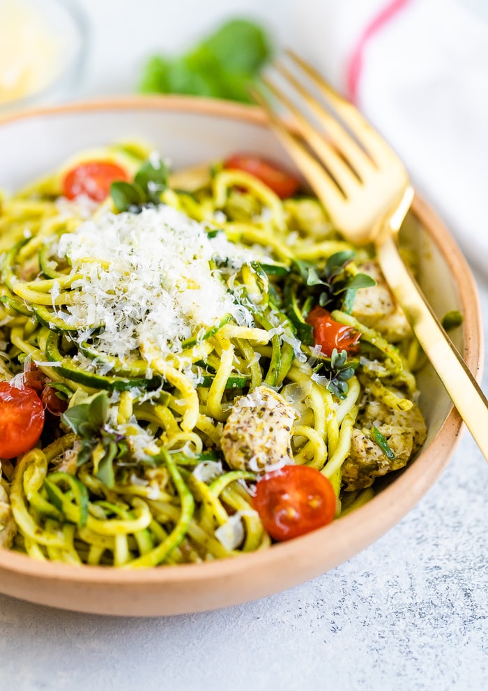 Bowl with pesto zucchini noodles with parmesan, chicken and tomatoes. Gold fork is resting on the side of the bowl.