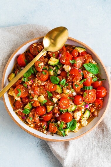 Serving bowl filled with a roasted tomato greek salad with a gold serving spoon resting on the bowl.