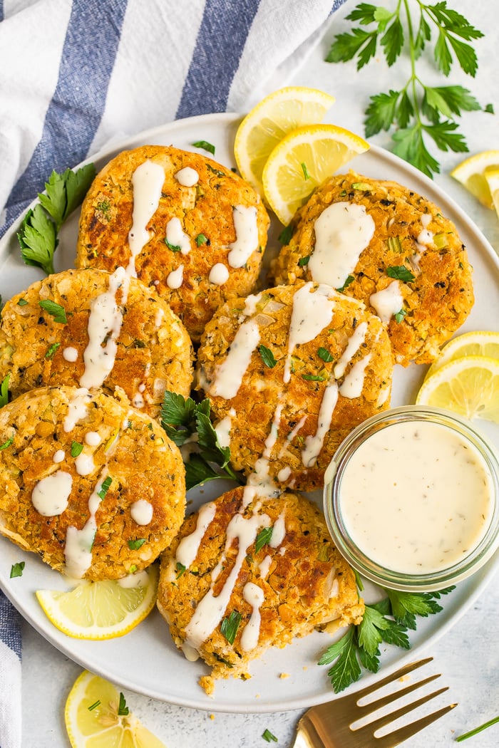 Photo aérienne d'une assiette avec 6 gâteaux de crabe végétaliens garnis de citron, de persil et d'un filet de sauce crémeuse.