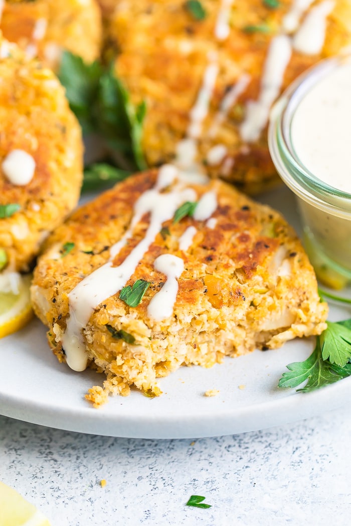 Close up photo of a vegan crab cake with a bite taken out of it on a plate. Cake is drizzled with a creamy dressing.