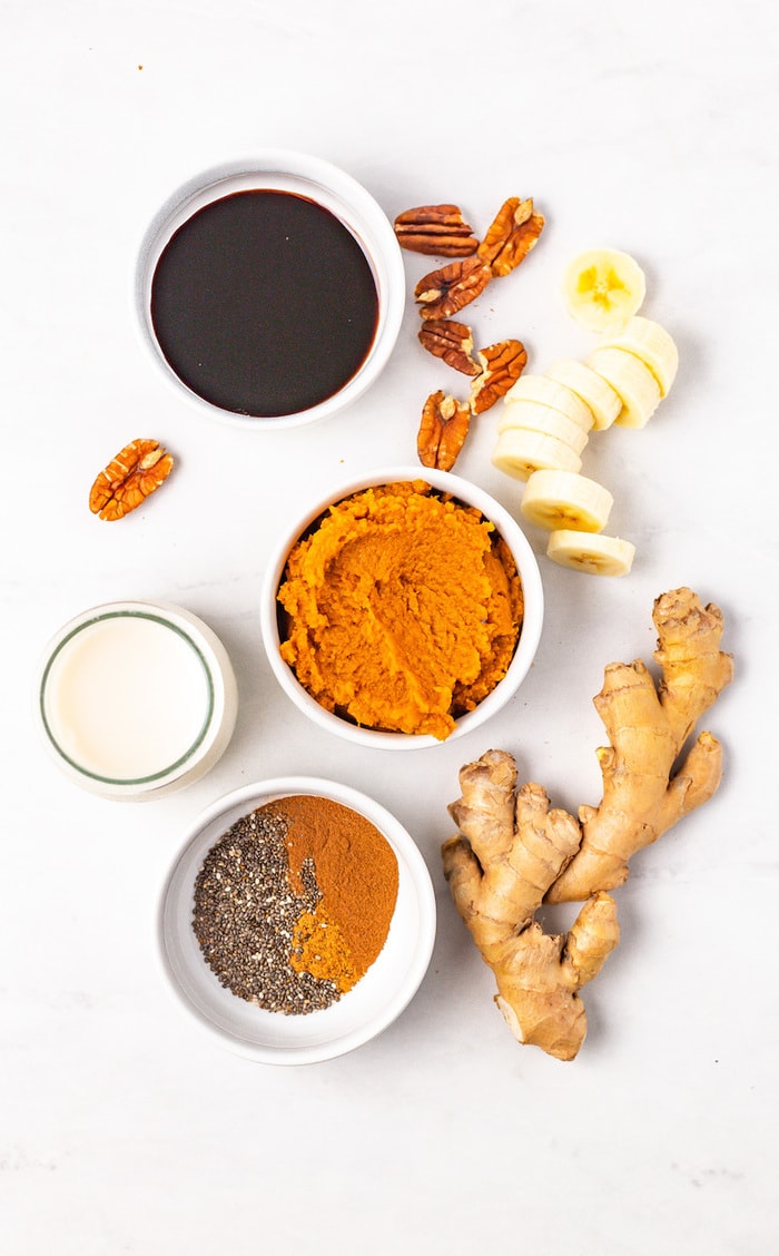 Molasses, almond milk, pumpkin, chia seeds, spices, ginger root, banana and pecans on a table in bowls measured out.