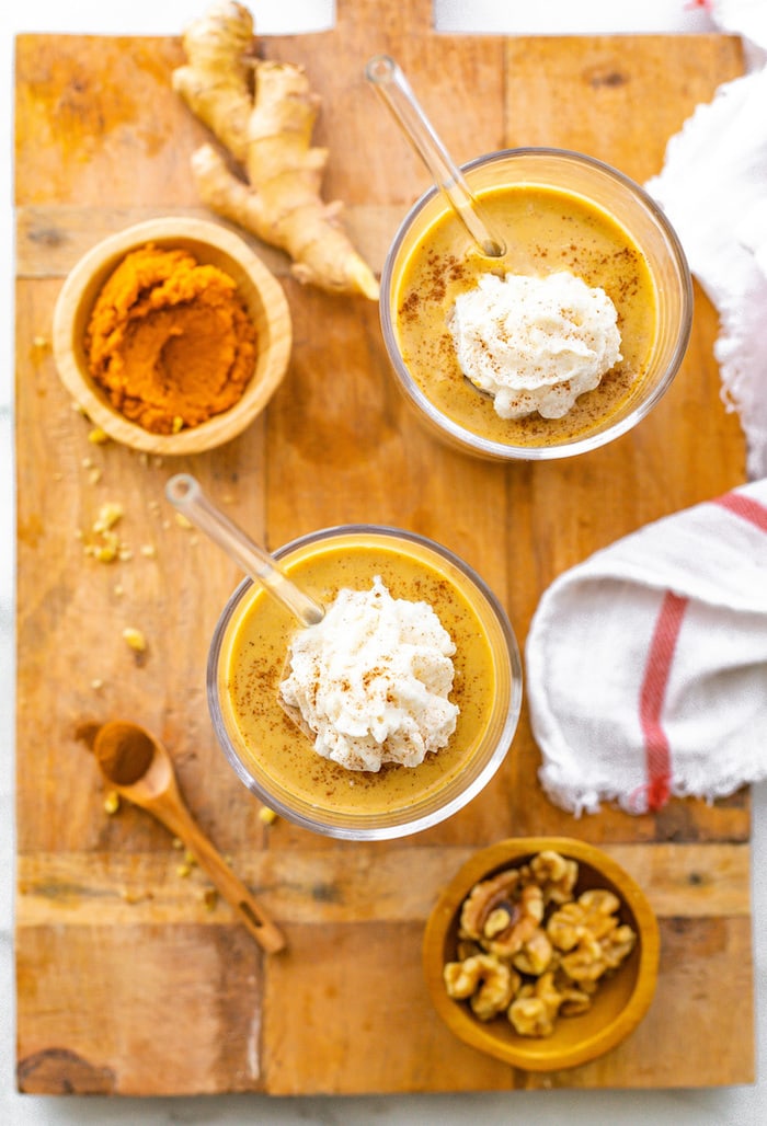 Bird's eye view photo of two pumpkin gingerbread smoothies topped with whipped cream and cinnamon. The smoothies are on a wooden board with spices, ginger, pumpkin and nuts are on the board.