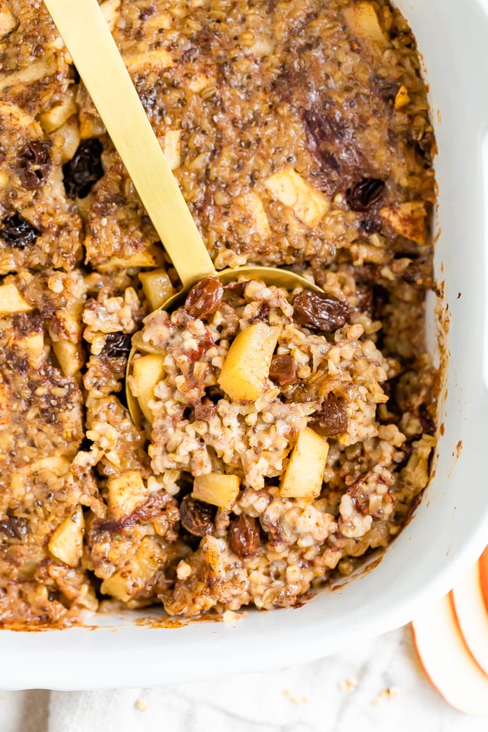 Overhead shot of apple cinnamon baked steel cut oatmeal in a white baking dish with a gold spoon scooping out a portion.