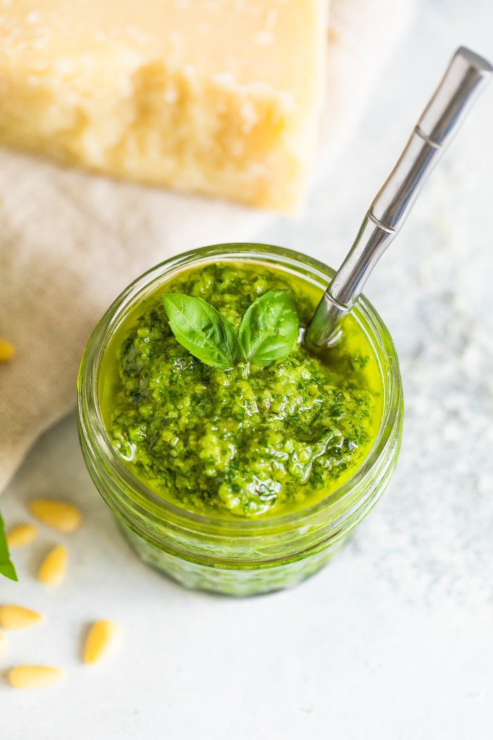 Overhead photo of a glass jar full of homemade pesto with a silver spoon in the jar. Pesto is topped with a couple sprigs of fresh basil.