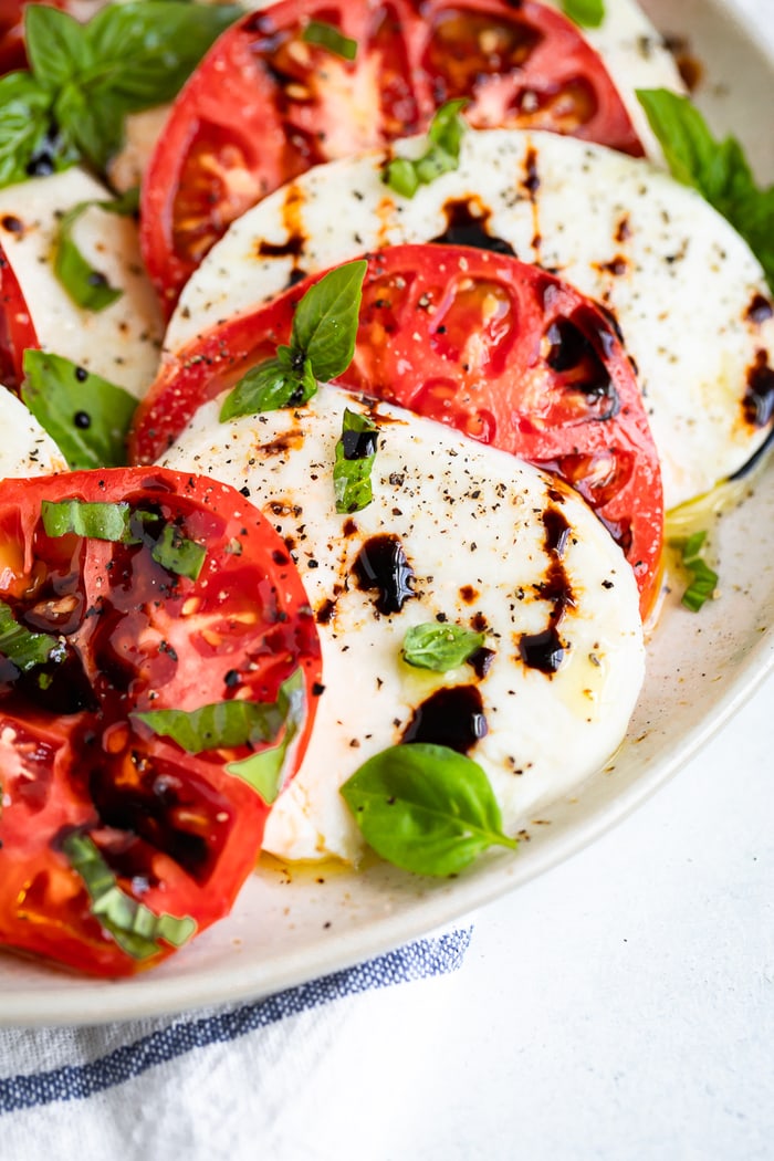 Close up photo of thick slices of tomato and mozzarella in a caprese salad topped with balsamic, pepper and basil.