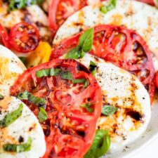 Close up photo of a caprese salad, slices of tomato and fresh mozzarella topped with balsamic, pepper and basil.