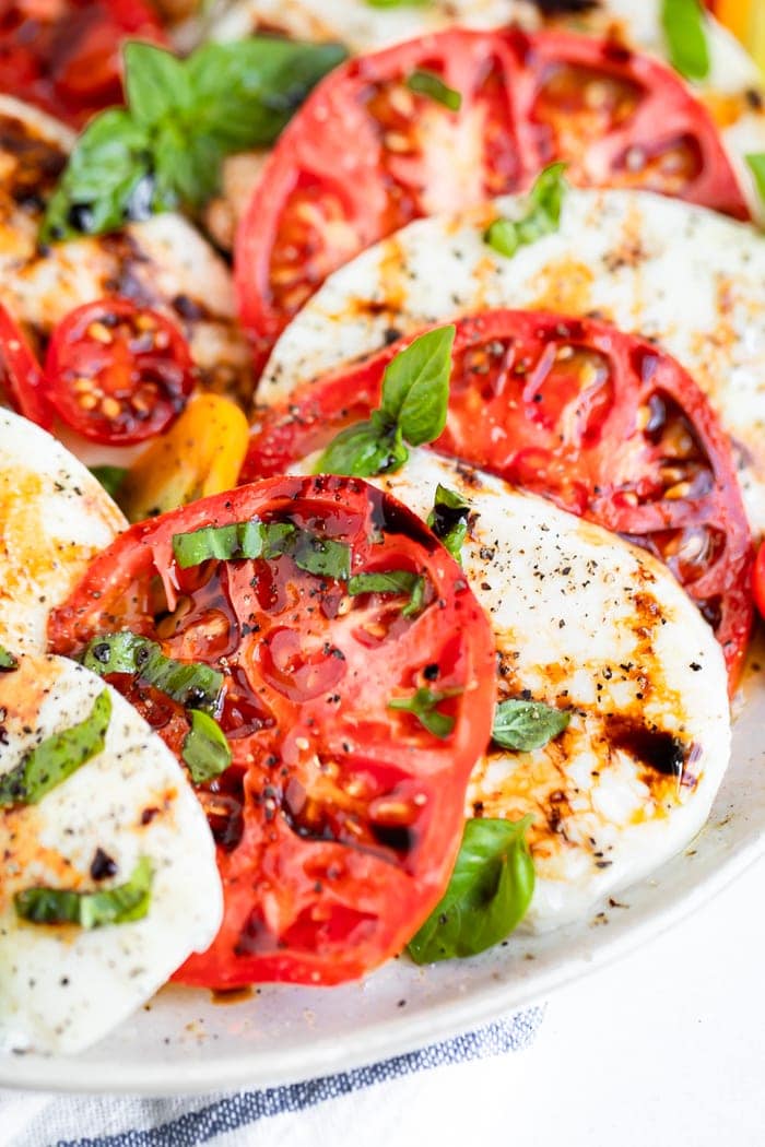 Close up photo of a caprese salad, slices of tomato and fresh mozzarella topped with balsamic, pepper and basil.