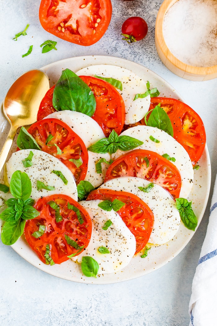 Thick slices of tomato and fresh mozzarella on a plate with a gold spoon and topped with pepper and fresh basil.