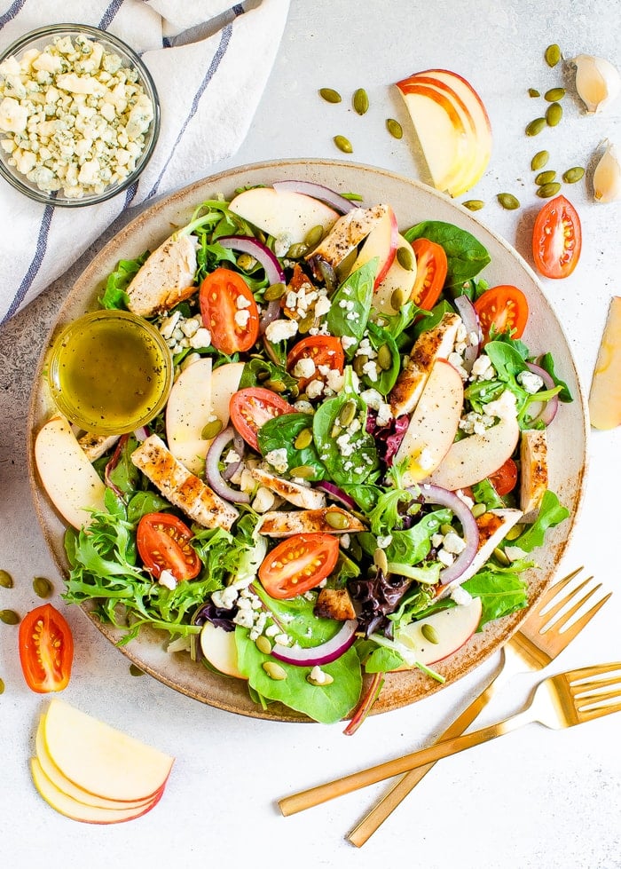 Plate with a fuji apple chicken salad. Two forks, pepitas, apples, tomatoes and gorgonzola are sprinkled around the plate.