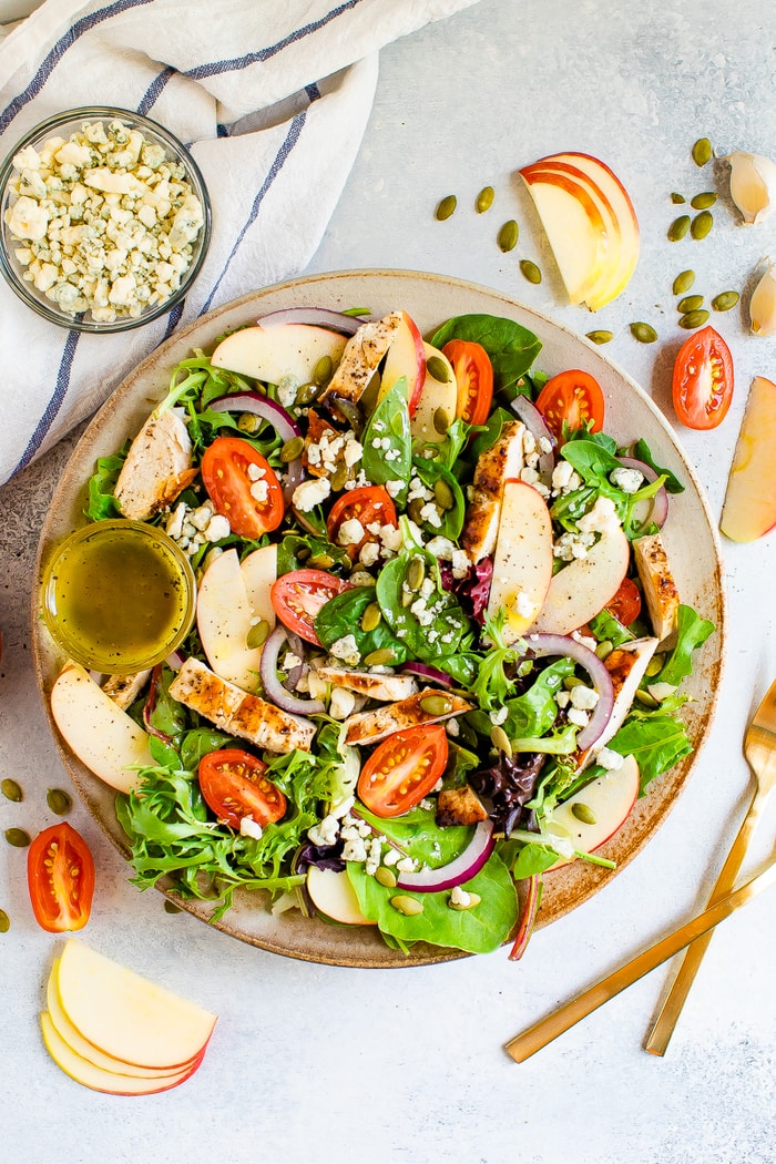 Plate with a fuji apple chicken salad. Two forks, pepitas, apples, tomatoes and gorgonzola are sprinkled around the plate.