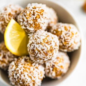 Bowl full of lemon energy balls rolled in coconut flakes. Slice of lemon is in the bowl.