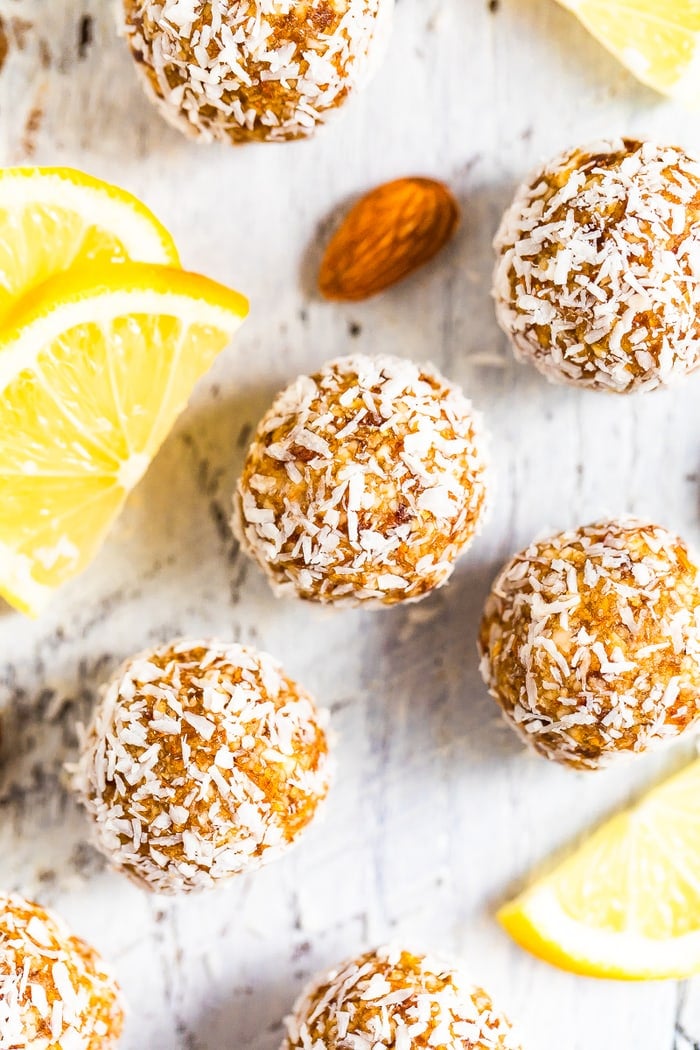 Lemon energy balls rolled in coconut flakes on a white wood table surrounded by lemon slices and almonds.