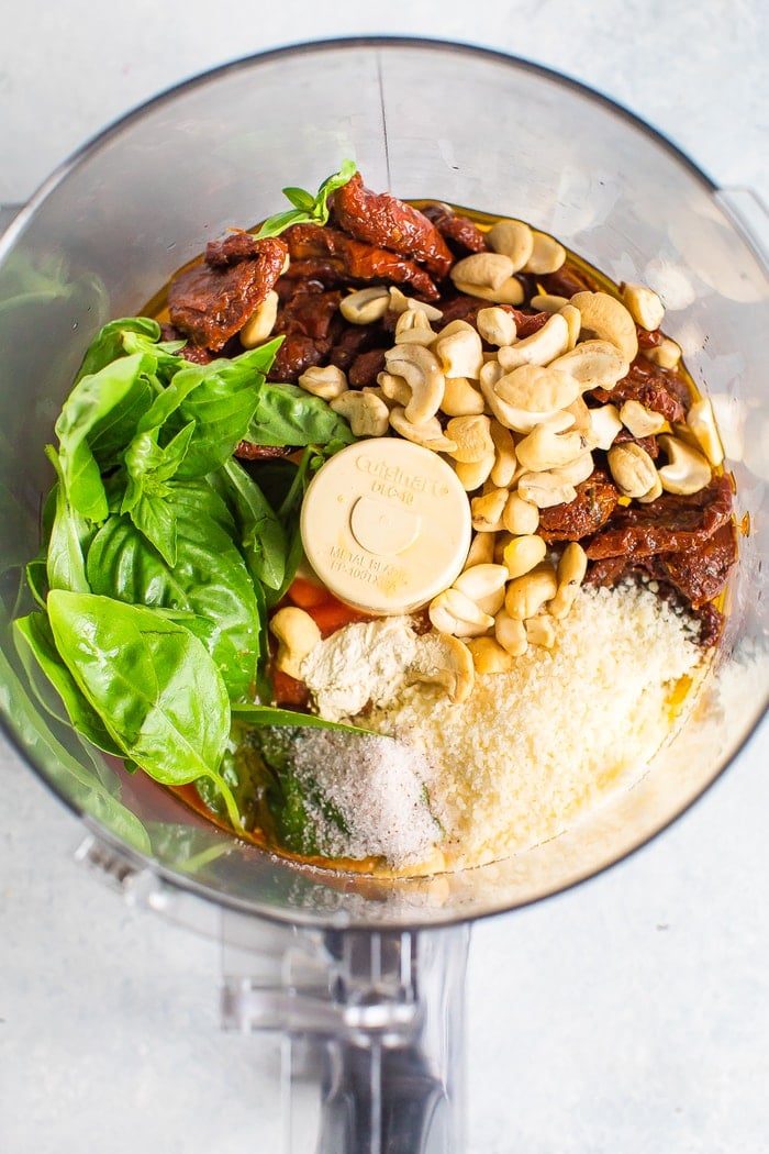 Food processor with the ingredients to make sun-dried tomato pesto-- basil, cashews, sun-dried tomatoes, parmesan and olive oil.