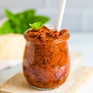 Glass jar on a wood cutting board full of sun-dried tomato pesto. The pesto has a white spoon stuck in it, and garnished with a fresh basil leaf.