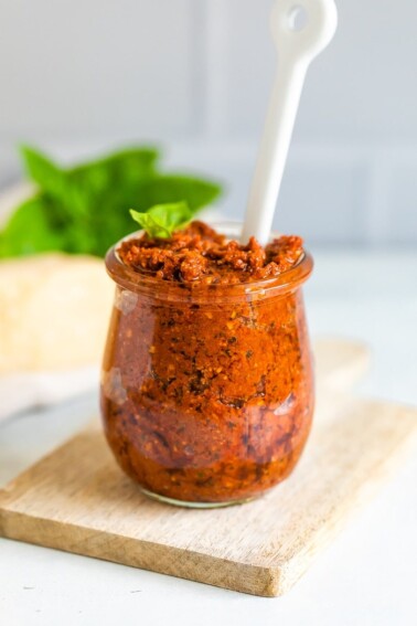 Glass jar on a wood cutting board full of sun-dried tomato pesto. The pesto has a white spoon stuck in it, and garnished with a fresh basil leaf.