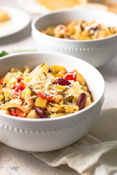 A white bowl with minestrone soup with shredded parmesan on top.