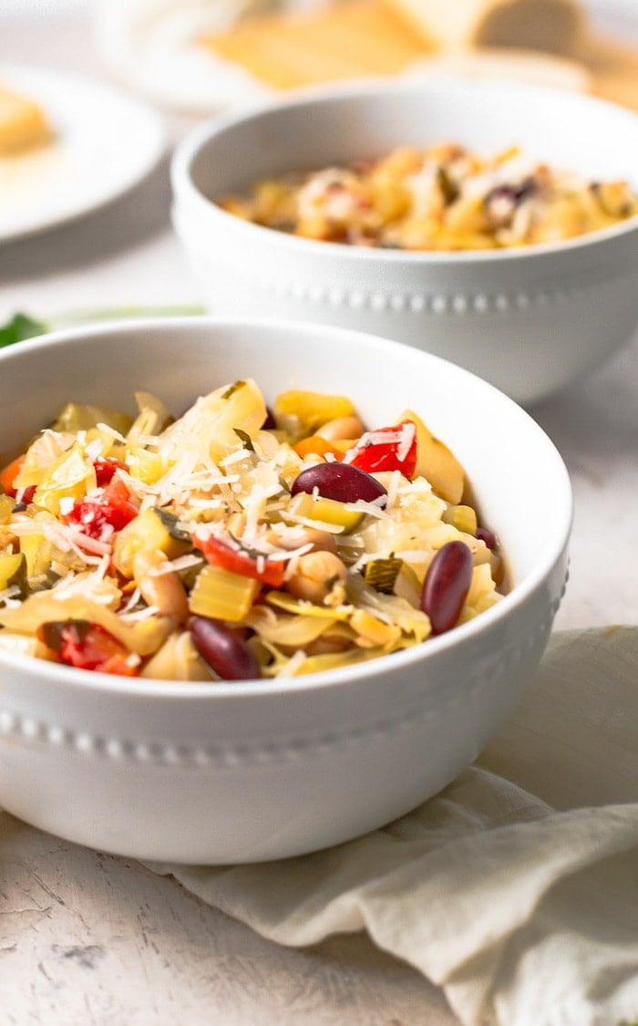 A white bowl with minestrone soup with shredded parmesan on top. 